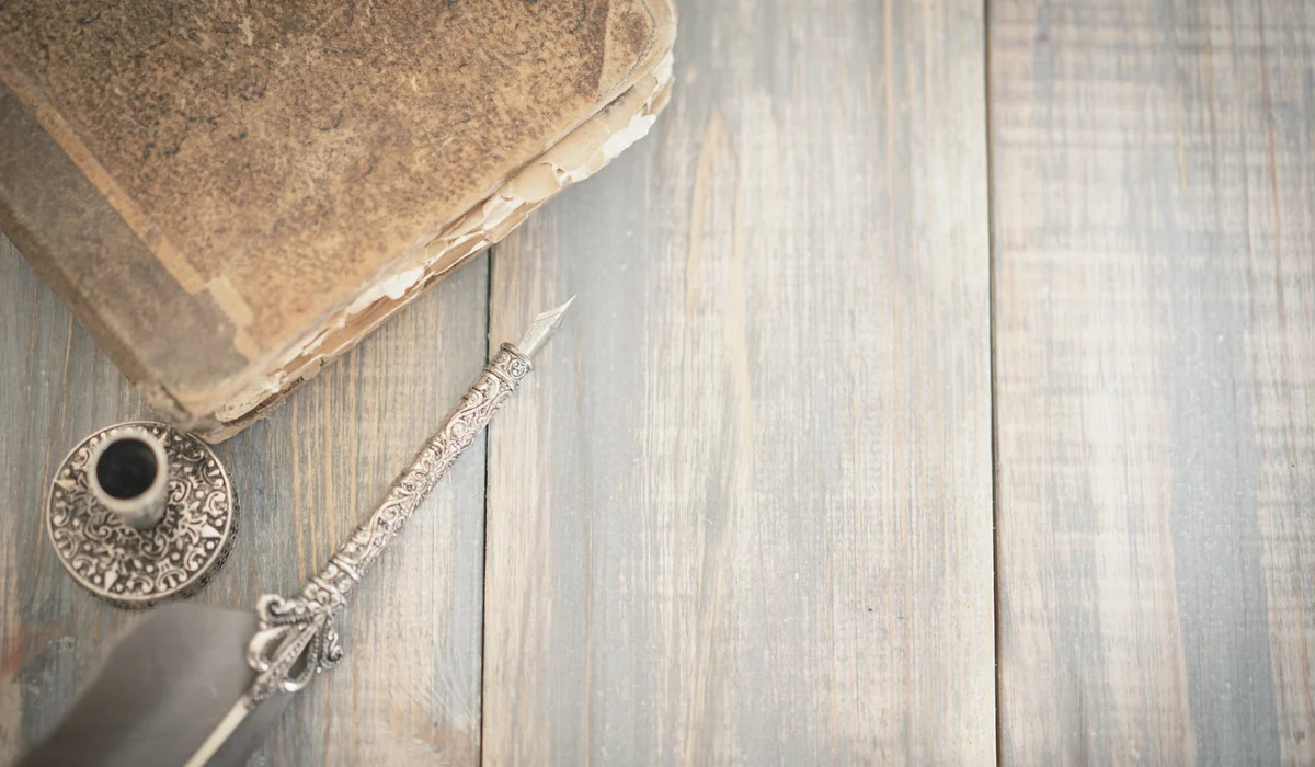 An overhead shot of a vintage book, ornate quill pen, and ink holder on a rustic wooden surface.