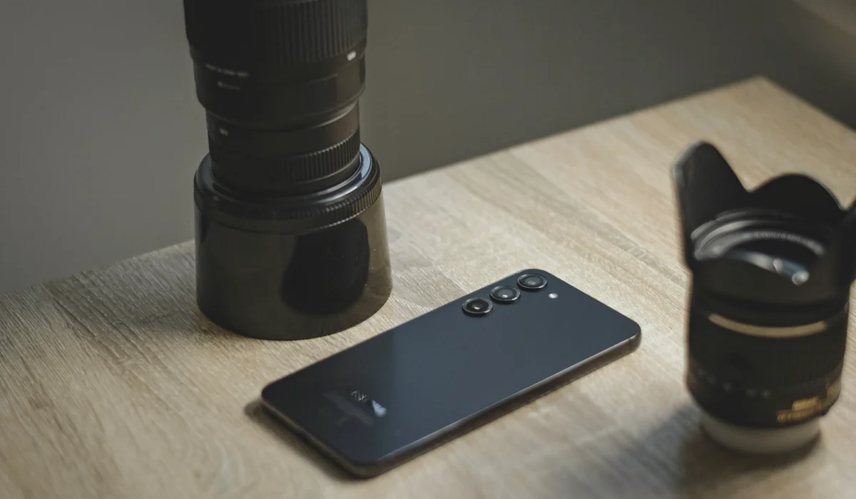 A sleek smartphone placed alongside camera lenses on a wooden desk, capturing technology and photography.