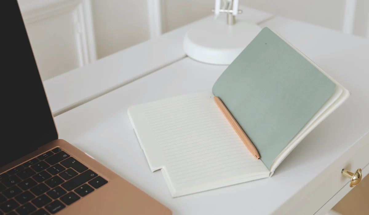 Minimalist workspace with an open notebook, pen, and laptop on a white desk.