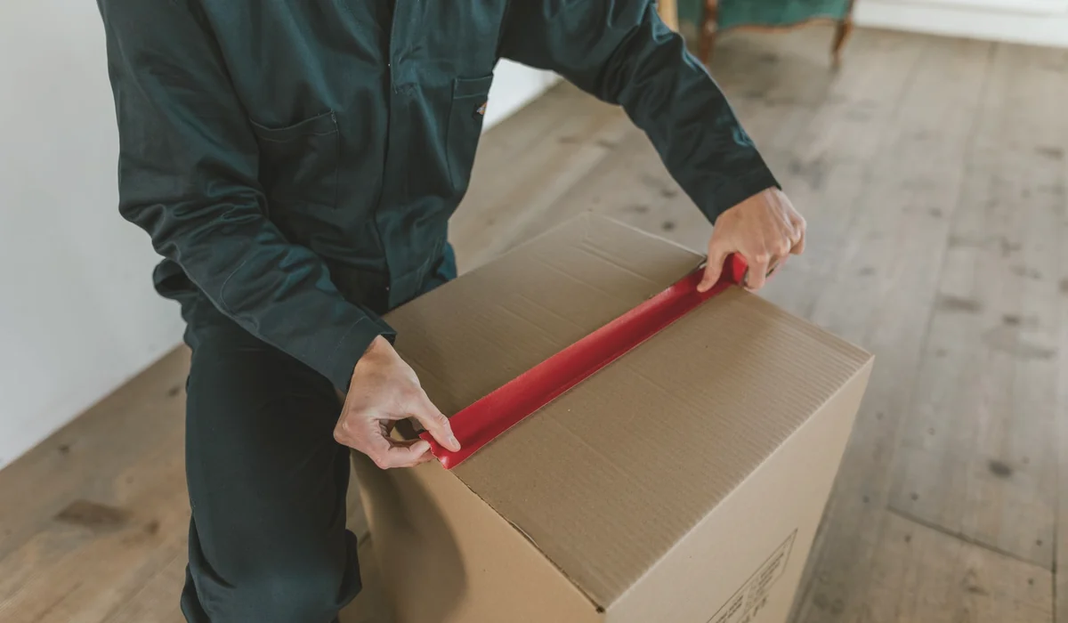Male courier sealing a cardboard box with red tape, indoor wooden floor setting.