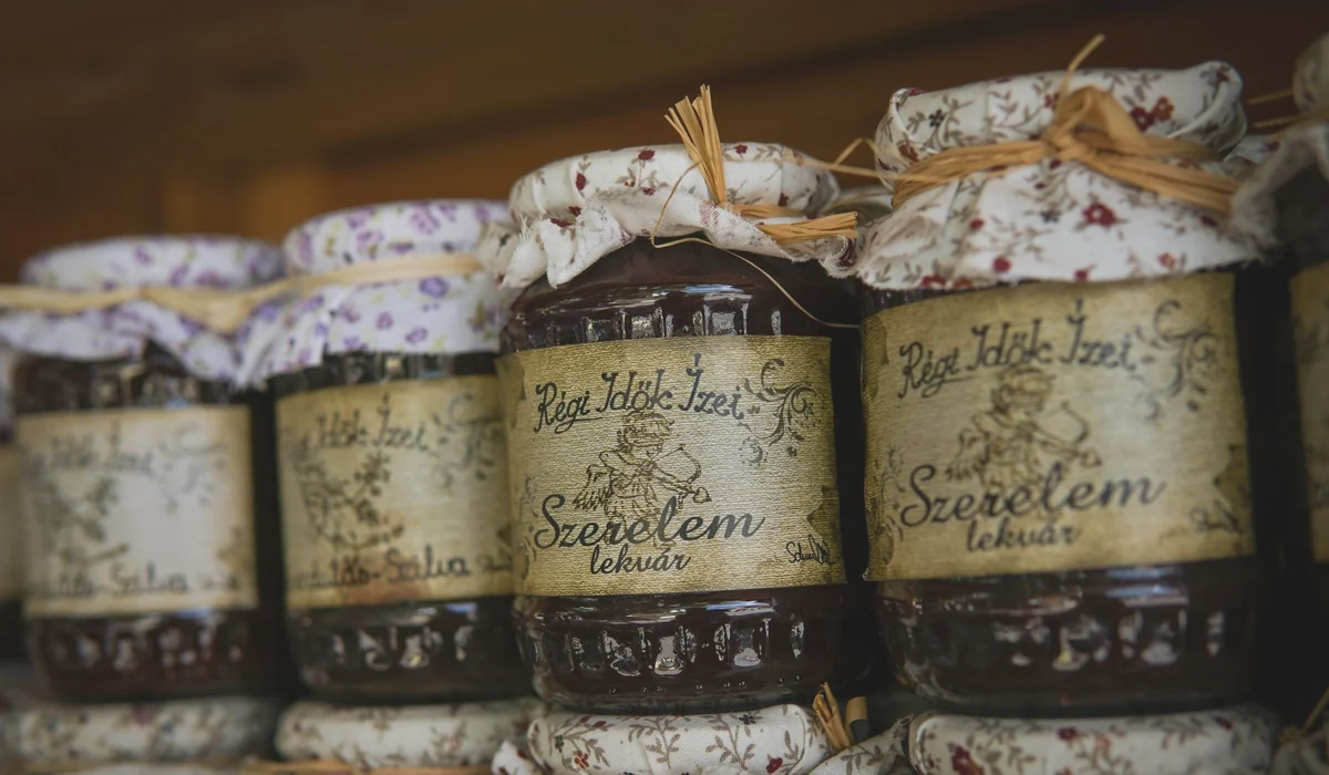 Close-up of rustic homemade jam jars with floral fabric covers and rustic labels on a wooden shelf.