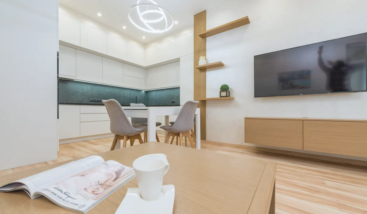 Interior of modern light room with table with mug and magazine in front of TV on wall next to chairs and open kitchen with cabinets and shelves