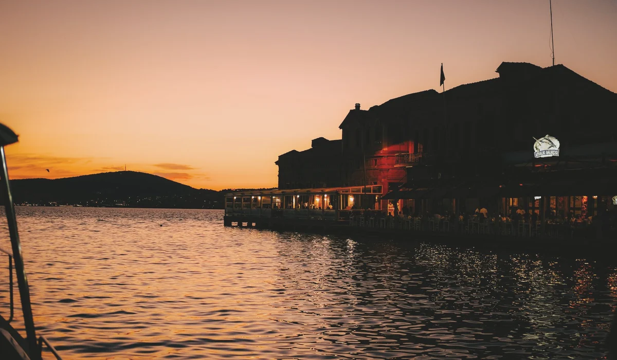 Silhouette of Ayvalık marina at dusk, with illuminated waterfront restaurants reflecting in the sea during a summer sunset.